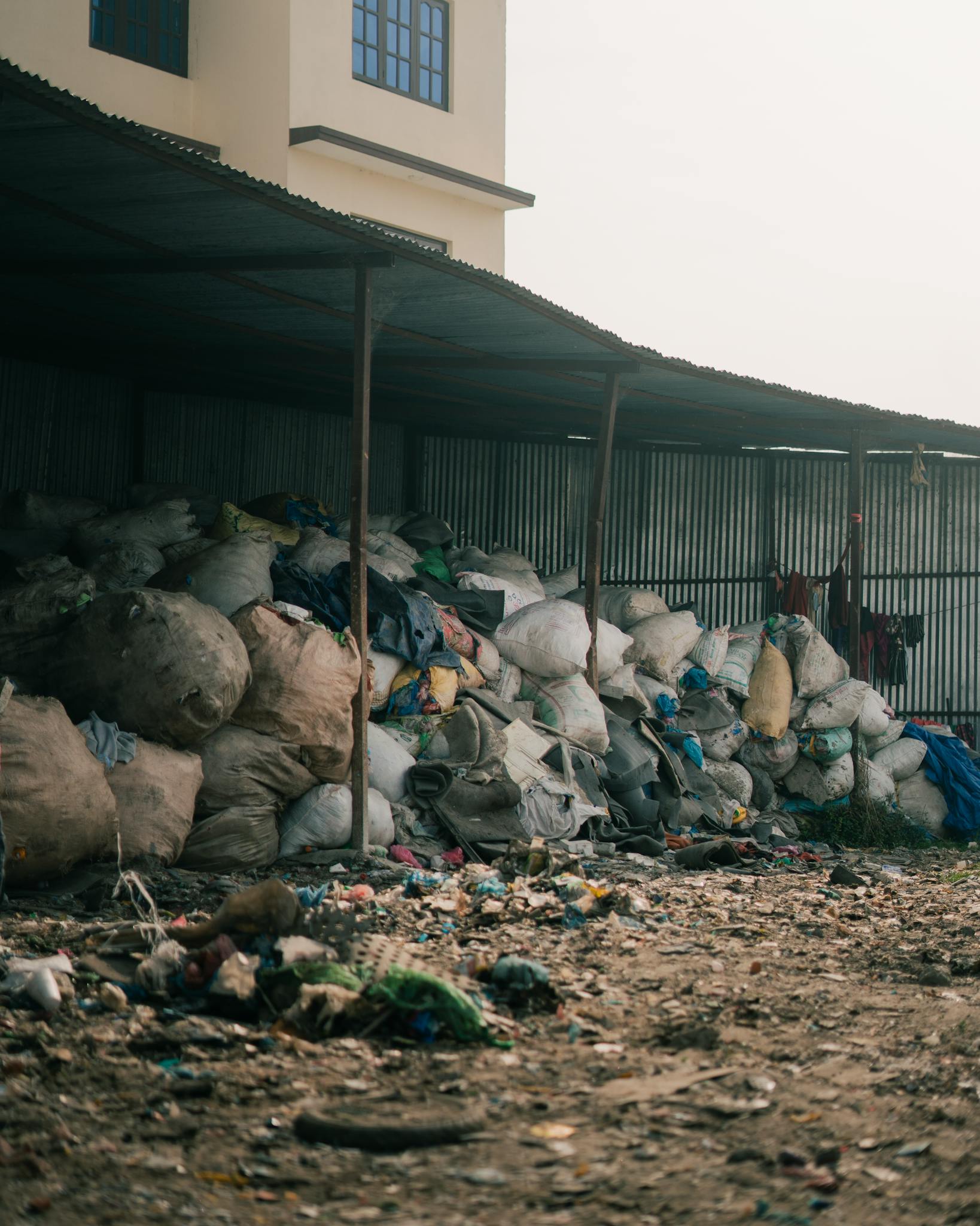 Piles of garbage bags stacked in an outdoor waste disposal area near industrial buildings.